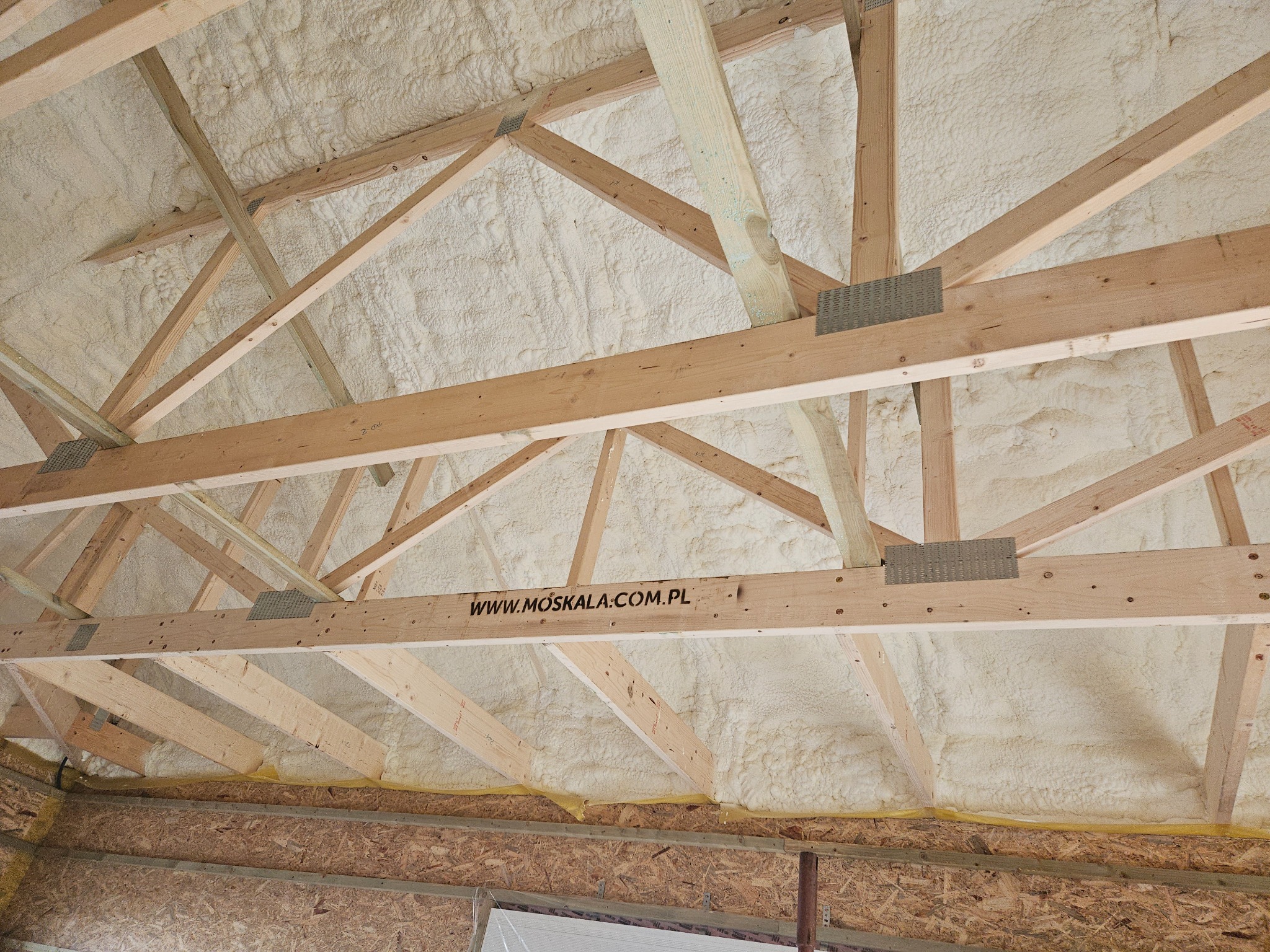 Poddasze z drewnianą konstrukcją pokryte grubą warstwą białej pianki izolacyjnej, widoczny adres strony internetowej na jednej z belek.