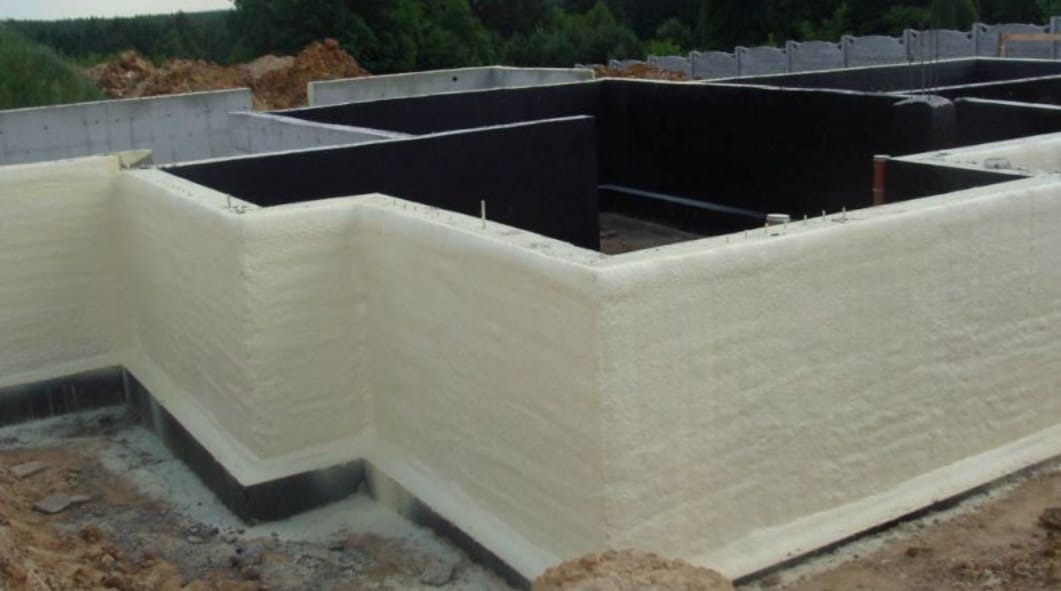 Fundamenty budynku zabezpieczone czarną hydroizolacją i pokryte warstwą jasnej pianki izolacyjnej, widoczne wystające pręty zbrojeniowe i teren budowy w tle.