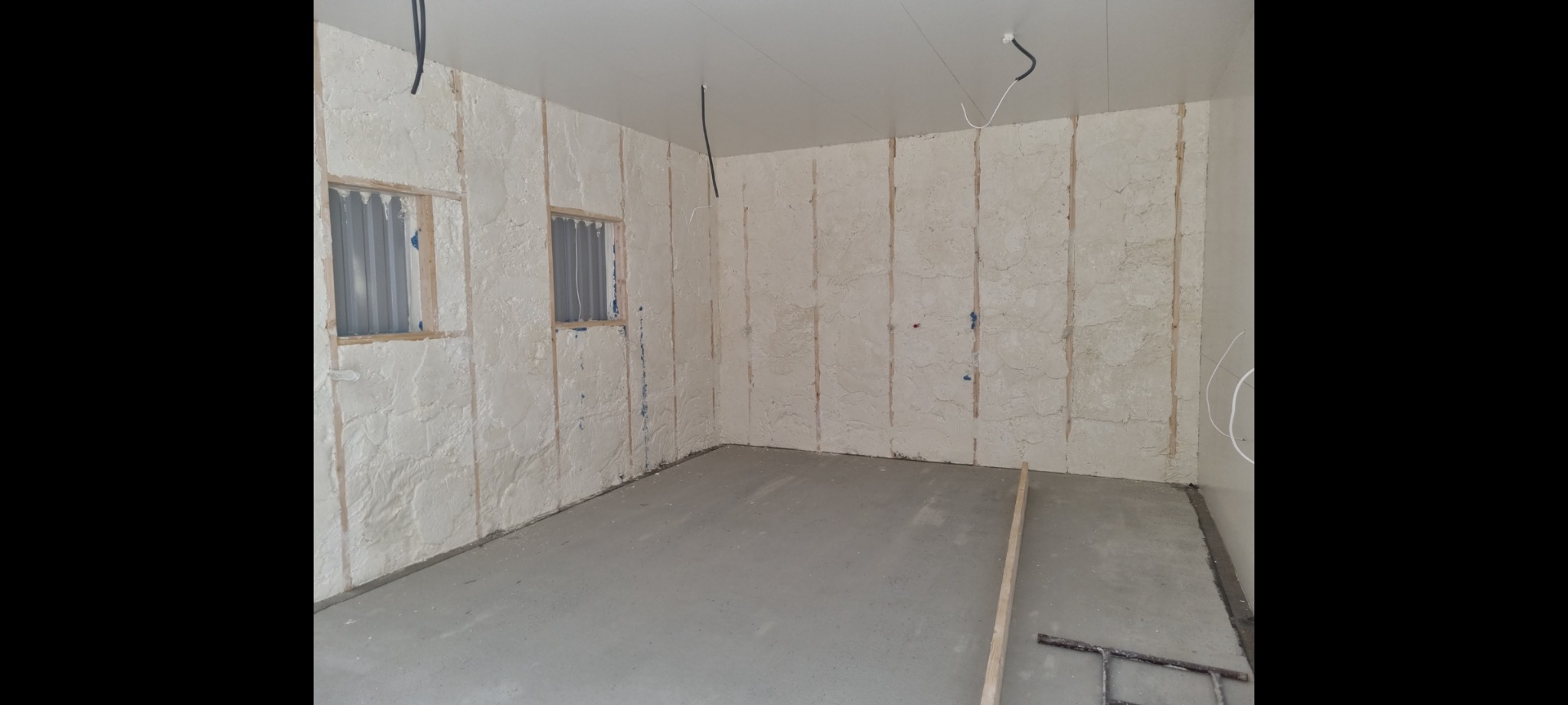 Wnętrze pomieszczenia po natrysku izolacji. Widoczne dwa okna zabezpieczone blachą falistą, ściany pokryte grubą warstwą izolacji natryskowej, świeża wylewka betonowa na podłodze i wystające...