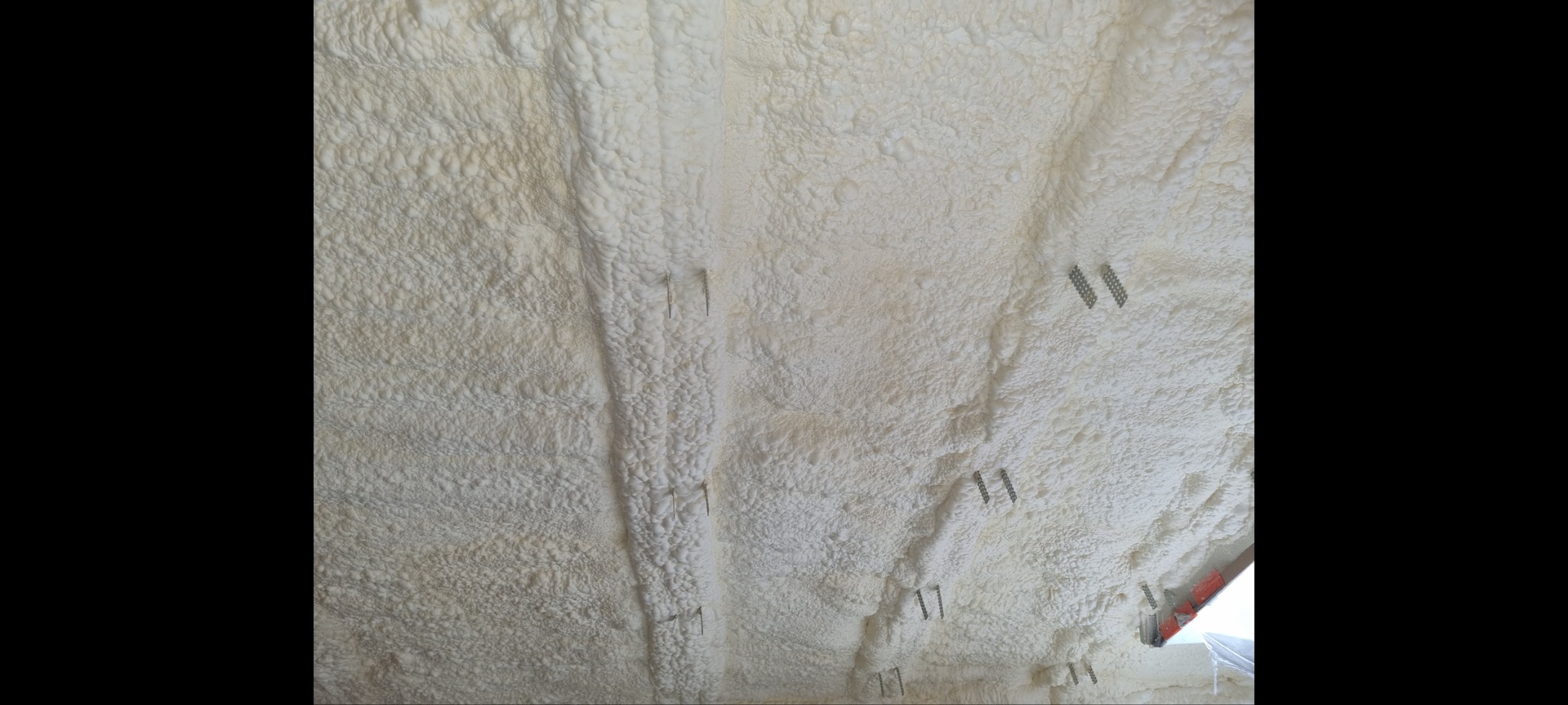 Beżowa, nierówna powierzchnia pokryta pianą izolacyjną z widocznymi metalowymi elementami mocującymi, tworząca wzór przypominający strukturę korala.