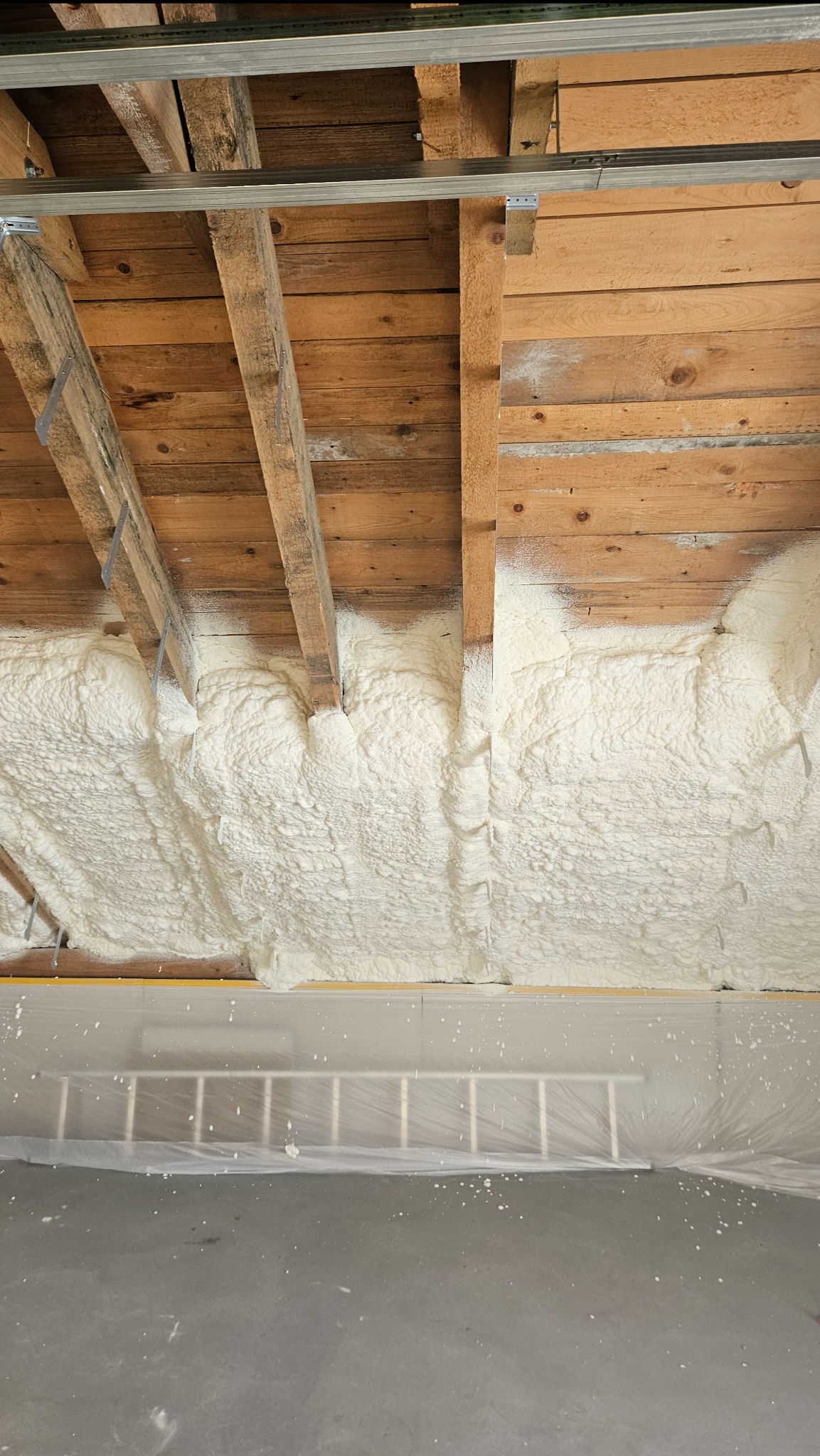 Poddasze z drewnianymi belkami i nałożoną warstwą białej piany izolacyjnej; w tle zabezpieczona folią drabina oparta o ścianę.