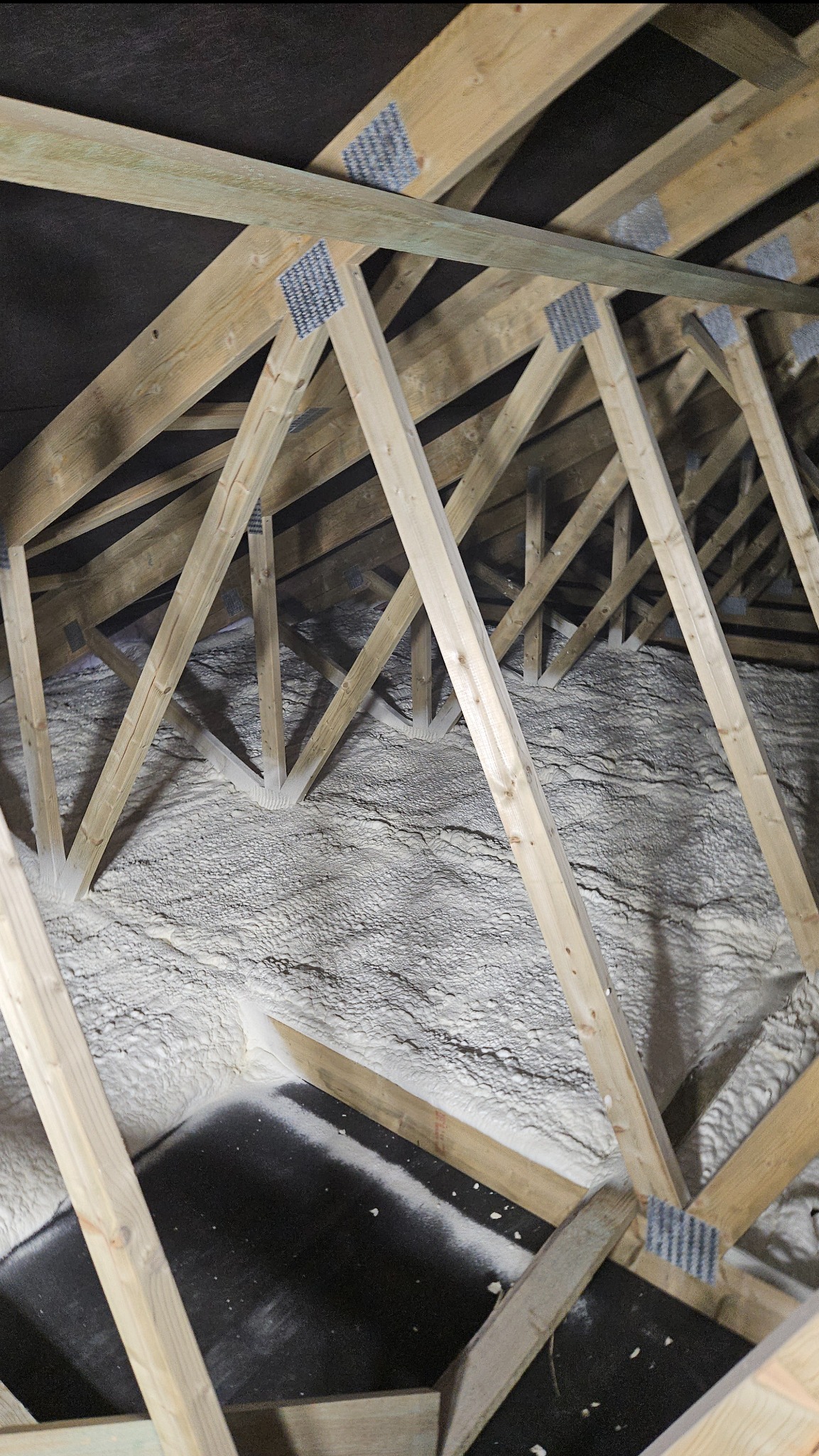 Poddasze z drewnianą konstrukcją krokwiową, pokryte białą, grubą warstwą izolacji natryskowej, widoczne fragmenty czarnej membrany dachowej.