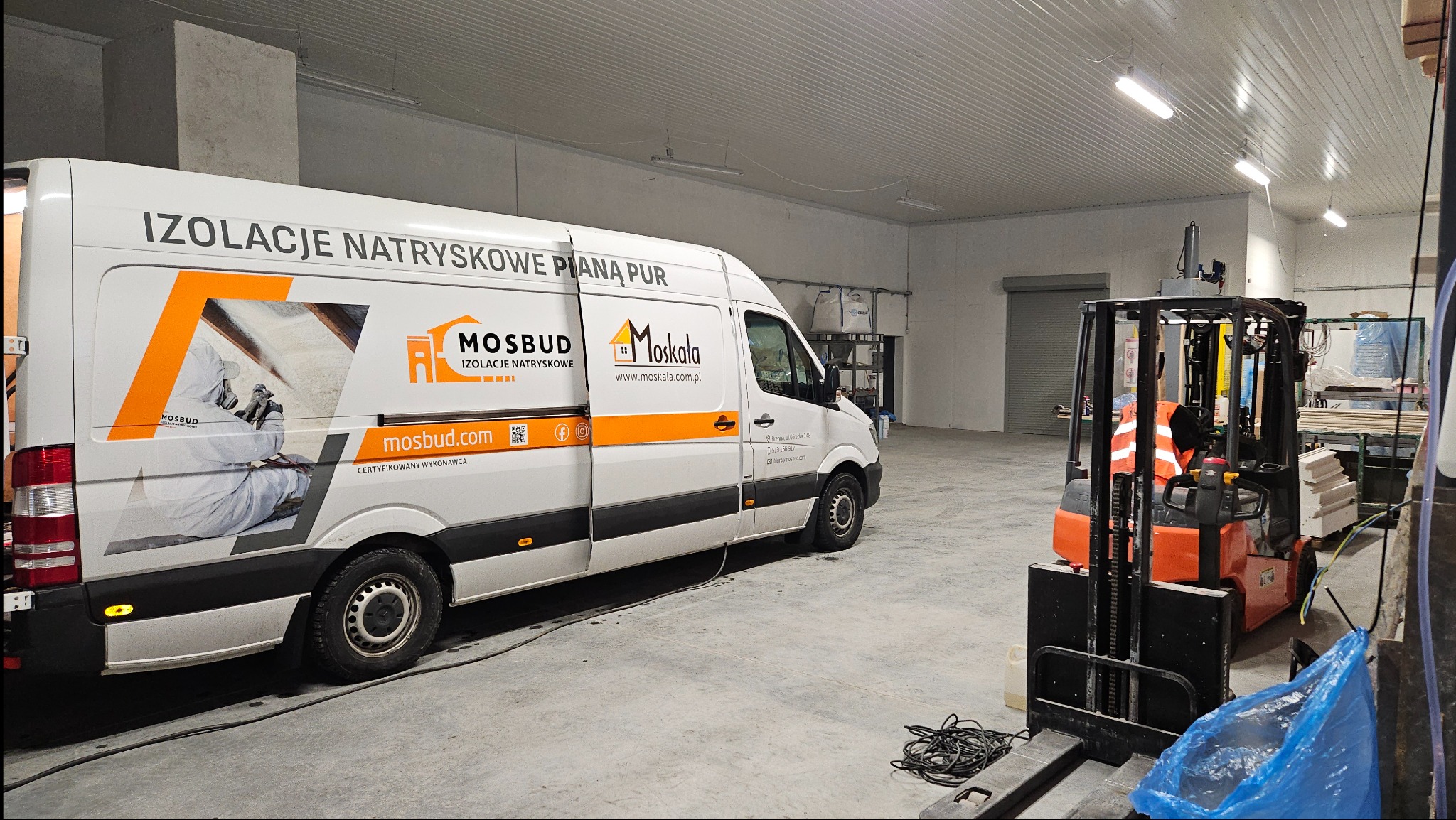 Biały samochód dostawczy z reklamą natryskowej piany PUR firm Mosbud i Moskala, zaparkowany w hali magazynowej obok wózka widłowego.