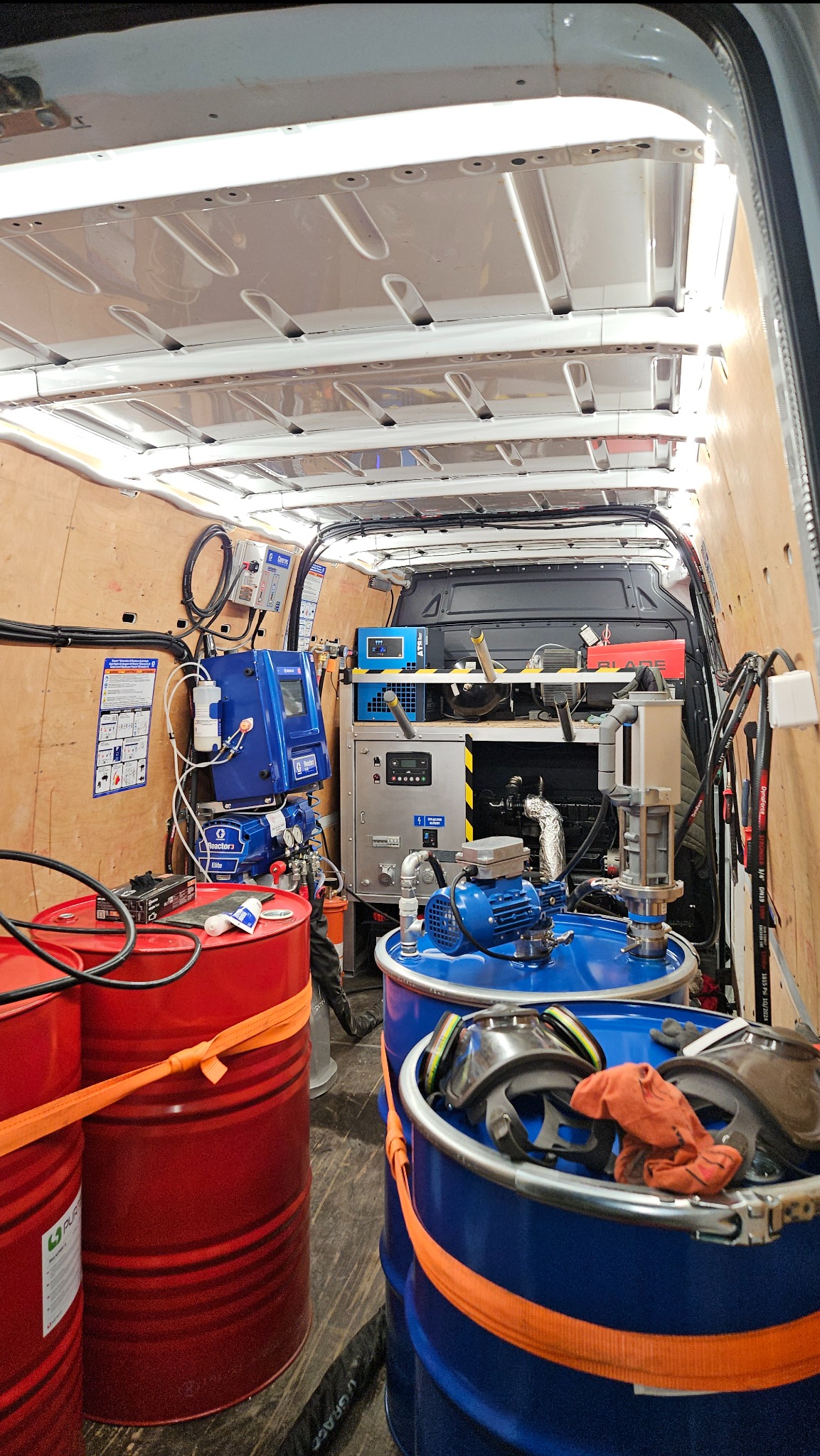Wnętrze furgonetki z urządzeniami do natrysku, w tym czerwone i niebieskie beczki z materiałami, maszyny natryskowe Graco Reactor Elite i akcesoria, widoczne elementy ochronne na beczce.