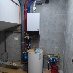 HPK Adrian Janczewski - Biały piec gazowy Viessmann zamontowany na szarej ścianie z płytkami, widoczne rury miedziane i plastikowe w kolorach czerwonym i niebieskim, zbiornik na wodę, filtr, podłoga zabezpieczona kartonem.