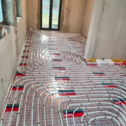 Instalacja ogrzewania podłogowego w surowym wnętrzu, widoczne białe rury na folii z czerwoną siatką, okno w tle.