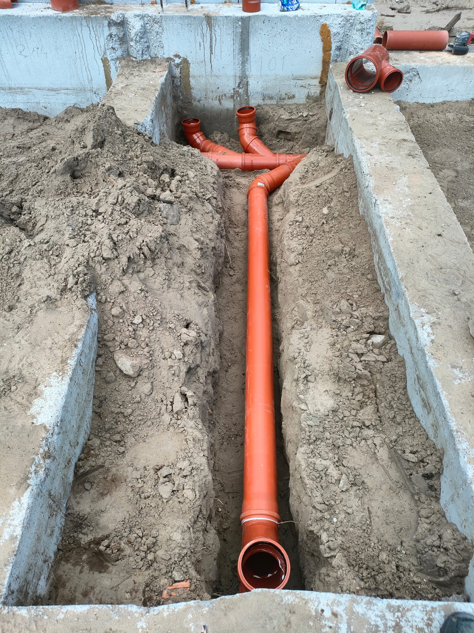 Pomarańczowe rury kanalizacyjne ułożone w wykopie ziemnym między betonowymi fundamentami budynku.