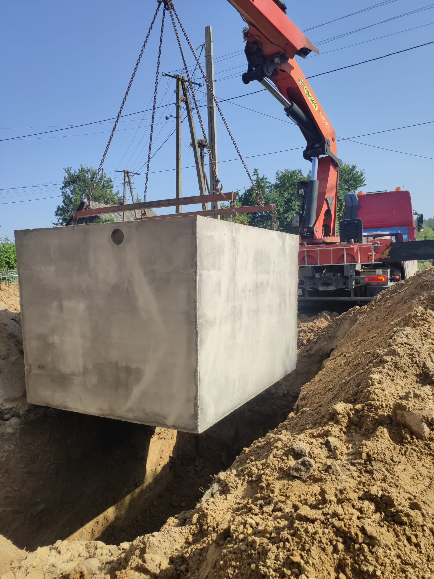 Ciężki betonowy zbiornik opuszczany dźwigiem do wykopu w ziemi, widoczne łańcuchy podtrzymujące element i ramię dźwigu. W tle słupy energetyczne i zieleń.
