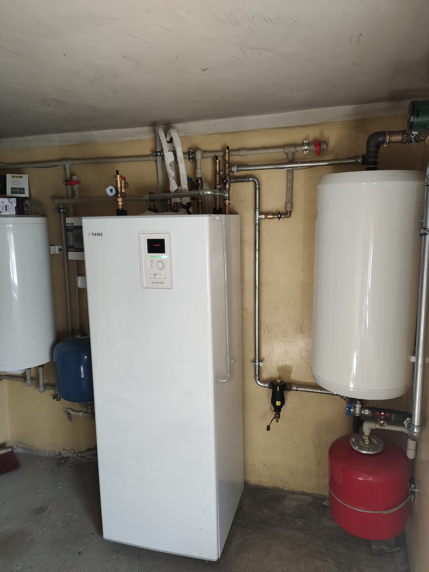 Biała pompa ciepła Nibe z podłączonymi rurami i zbiornikami w kotłowni, widoczny manometr i inne elementy instalacji grzewczej.