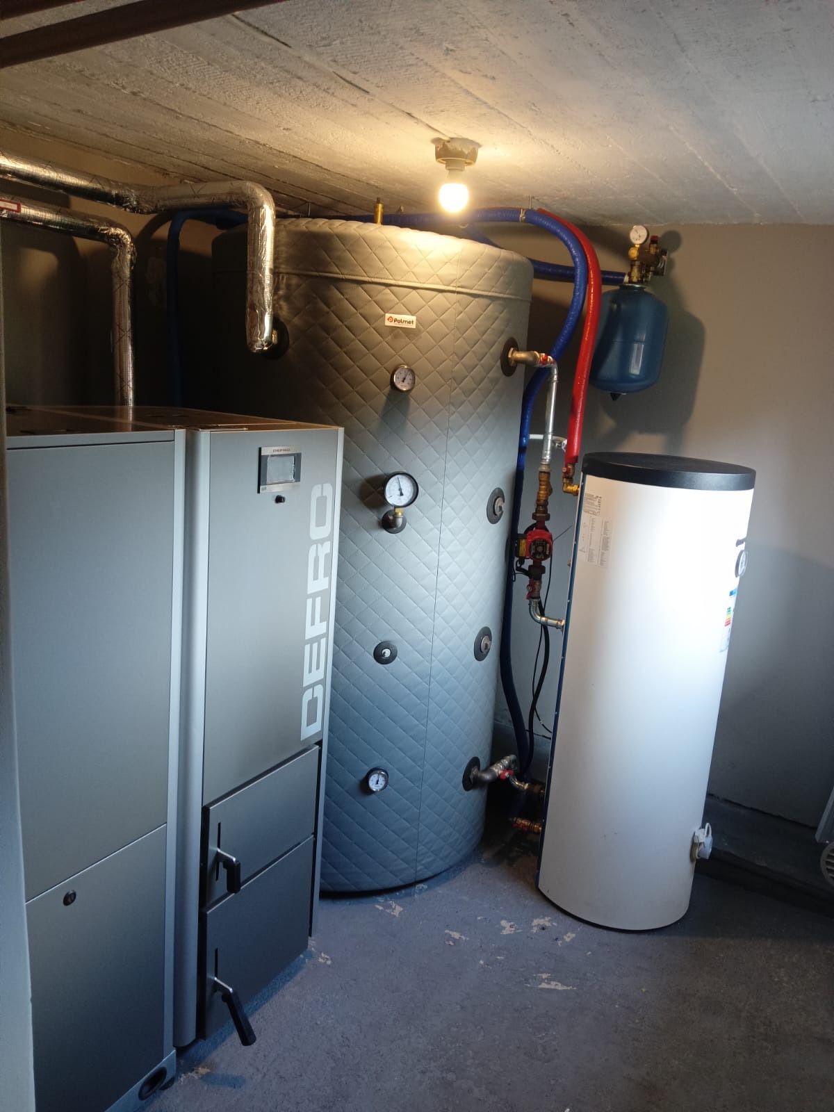 Kotłownia z kotłem Defro, zbiornikiem akumulacyjnym Palmet w pikowanej obudowie i białym zasobnikiem ciepłej wody, widoczne rury instalacji grzewczej i manometry.