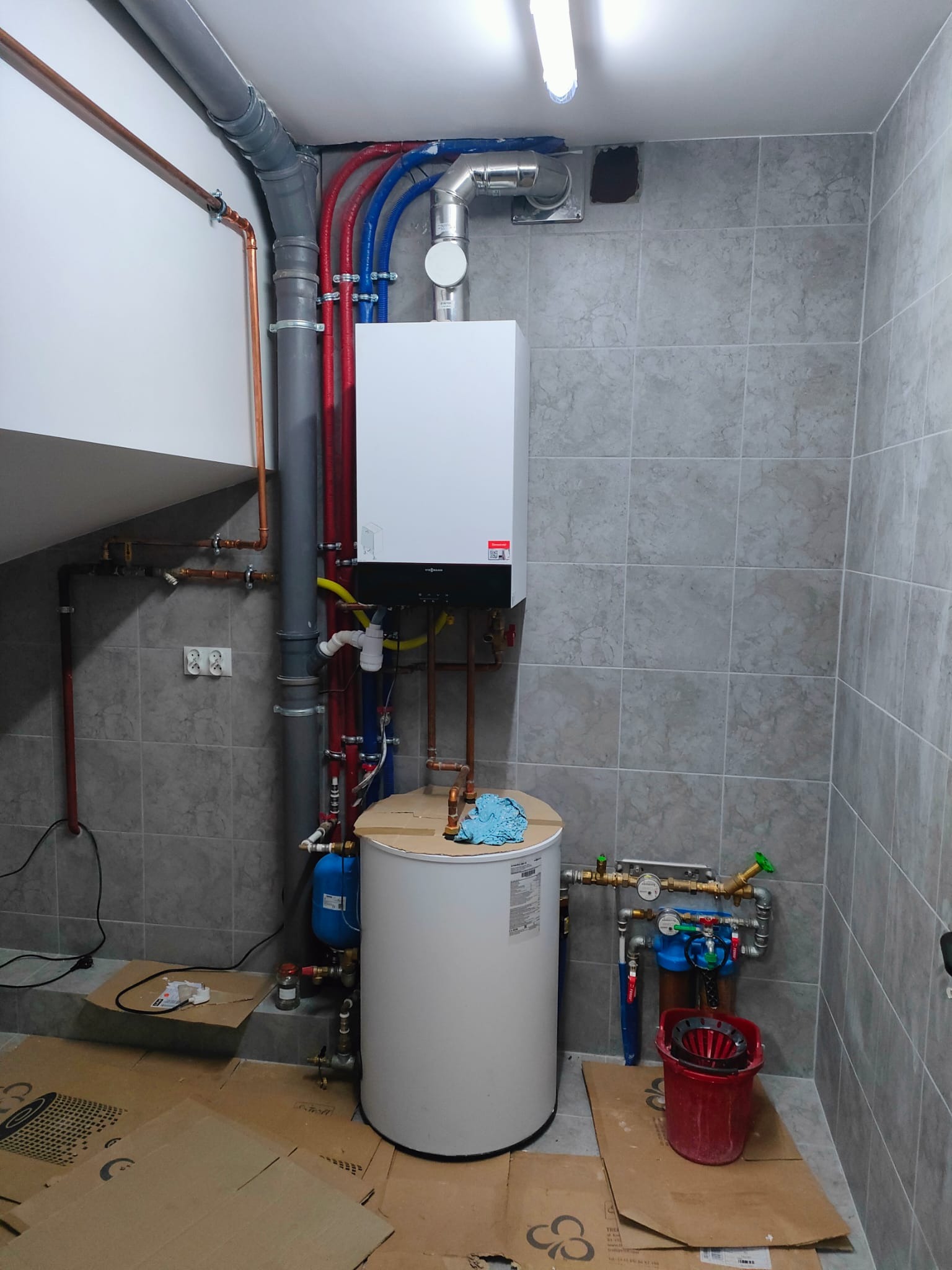 Biały piec gazowy Viessmann zamontowany na szarej ścianie z płytkami, widoczne rury miedziane i plastikowe w kolorach czerwonym i niebieskim, zbiornik na wodę, filtr, podłoga zabezpieczona kartonem.