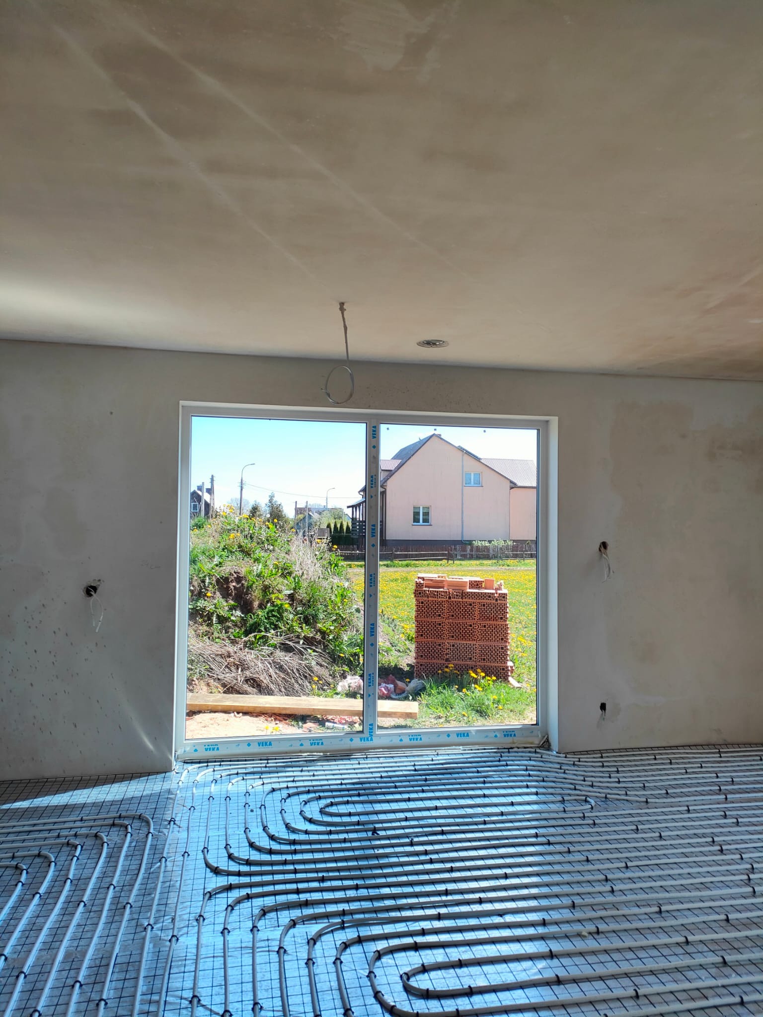 Wnętrze w stanie surowym z widoczną instalacją ogrzewania podłogowego, rurki ułożone na siatce, okno z widokiem na ogród i stertę cegieł.