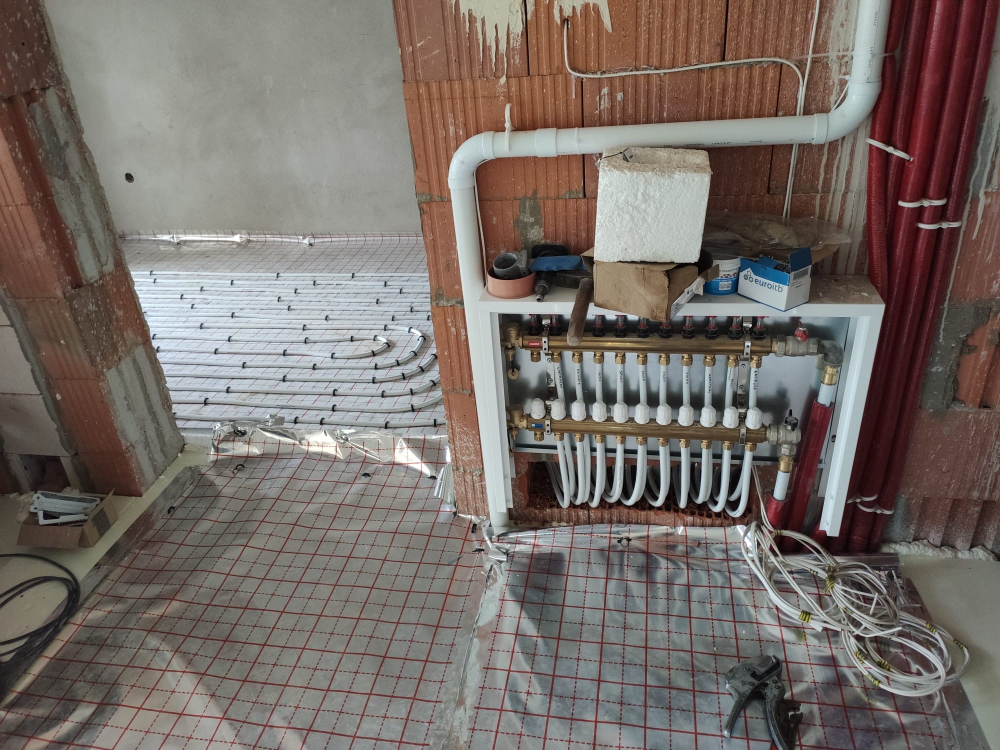 Rozdzielacz ogrzewania podłogowego z rurami, narzędziami i materiałami budowlanymi w surowym wnętrzu, widok na ułożoną instalację ogrzewania podłogowego.