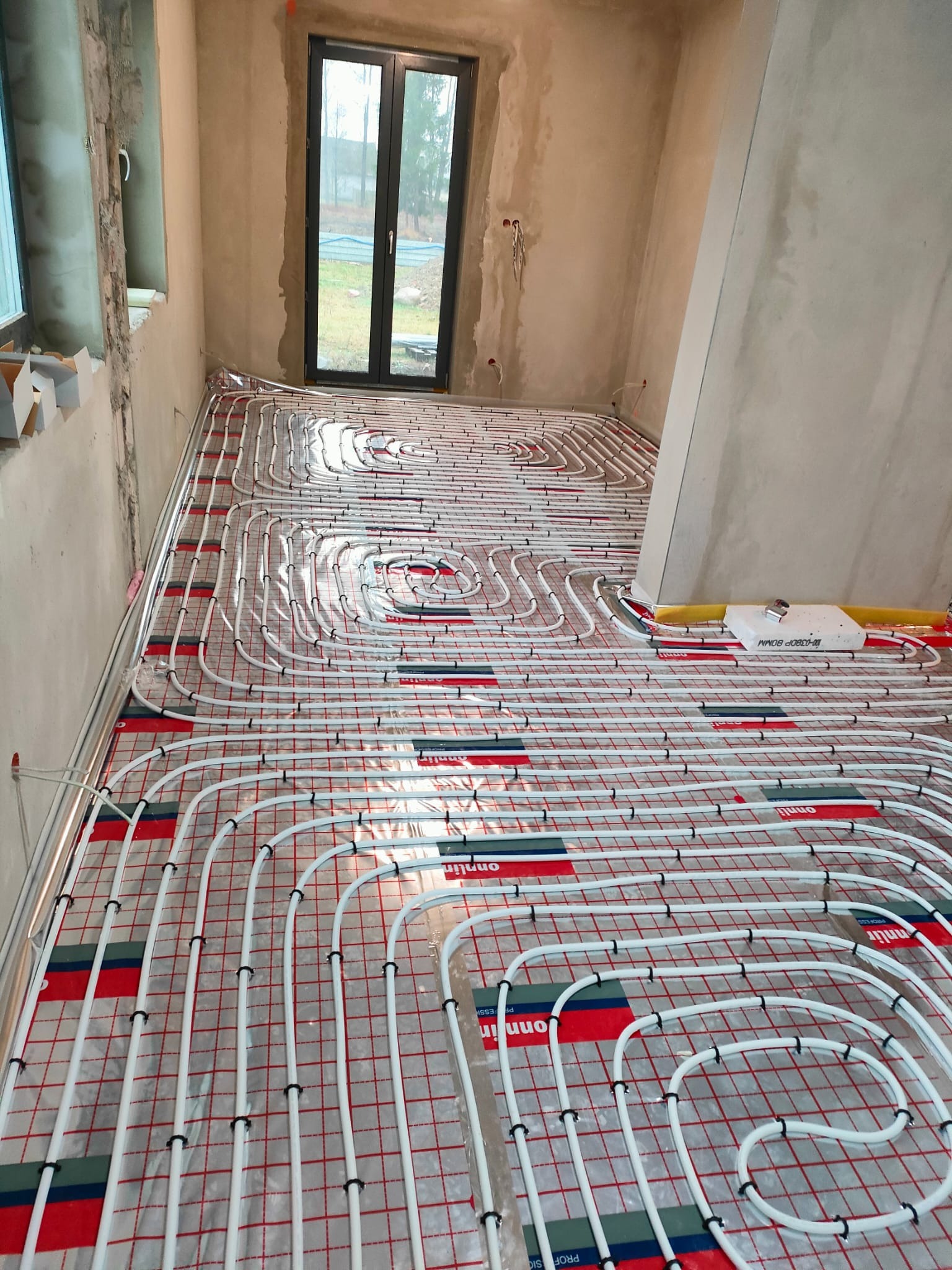 Instalacja ogrzewania podłogowego w surowym wnętrzu, widoczne białe rury na folii z czerwoną siatką, okno w tle.
