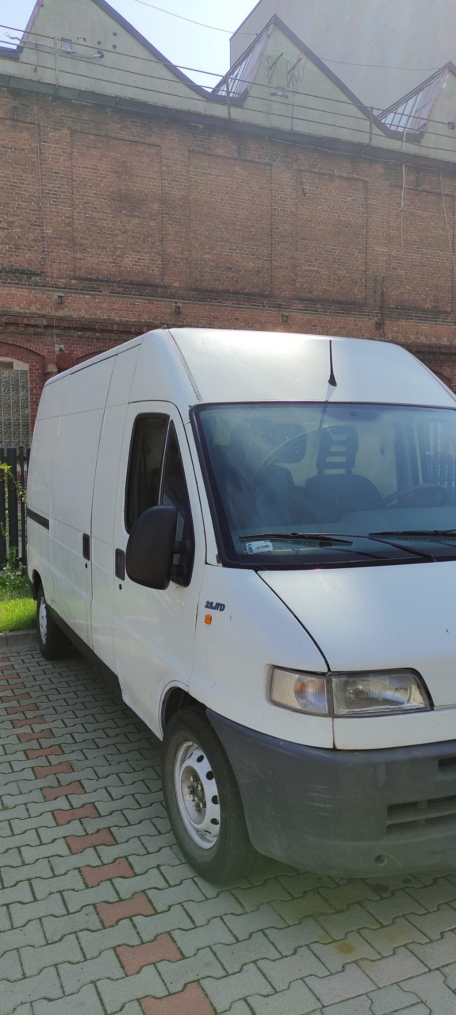 Biały samochód dostawczy 2.8 JTD zaparkowany na brukowanej nawierzchni z ceglanym budynkiem w tle.