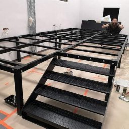 Usługi remontowo-budowlane Marcin Jakoniuk - Metalowa konstrukcja pod podłogę z schodami w surowym wnętrzu biura, pomarańczowa taśma na podłodze, mężczyzna siedzący na konstrukcji.