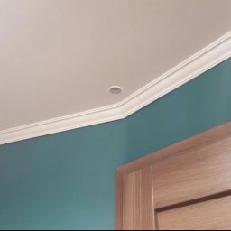 Usługi remontowo-budowlane Marcin Jakoniuk - Turkusowa ściana z białym sufitem z listwą dekoracyjną i wpuszczonymi punktami świetlnymi, fragment drewnianej ościeżnicy drzwiowej w prawym dolnym rogu.