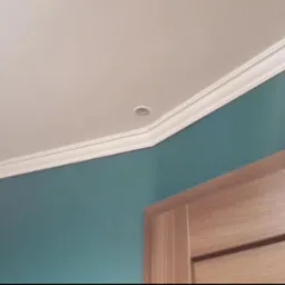 Turkusowa ściana z białym sufitem z listwą dekoracyjną i wpuszczonymi punktami świetlnymi, fragment drewnianej ościeżnicy drzwiowej w prawym dolnym rogu.