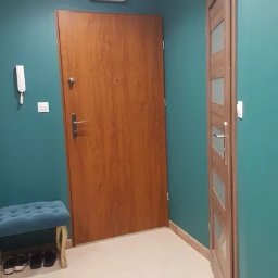 Usługi remontowo-budowlane Marcin Jakoniuk - Korytarz w odcieniach turkusu z dwiema drewnianymi drzwiami: wejściowymi i wewnętrznymi z przeszkleniami, na podłodze jasne płytki, mała pufa z butami pod ścianą.