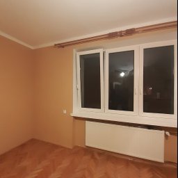 Usługi remontowo-budowlane Marcin Jakoniuk - Pomalowane na ciepły odcień wnętrze pokoju z parkietem ułożonym w jodełkę, białym grzejnikiem pod trzyskrzydłowym oknem z otwartym jednym skrzydłem.