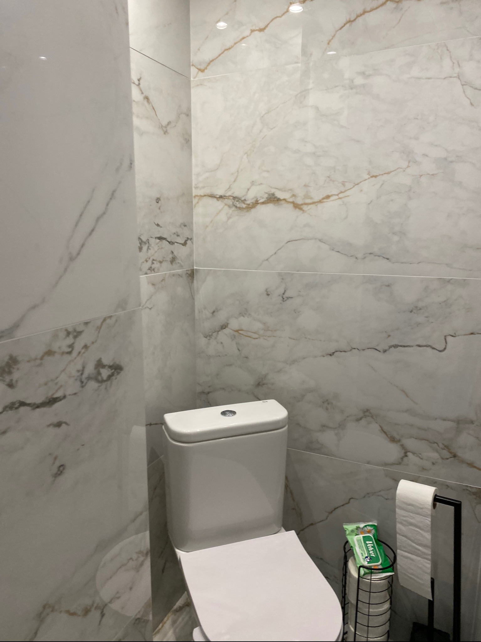 Nowoczesna toaleta z białymi płytkami imitującymi marmur, uzupełniona o stojak na papier toaletowy i zapas chusteczek higienicznych.