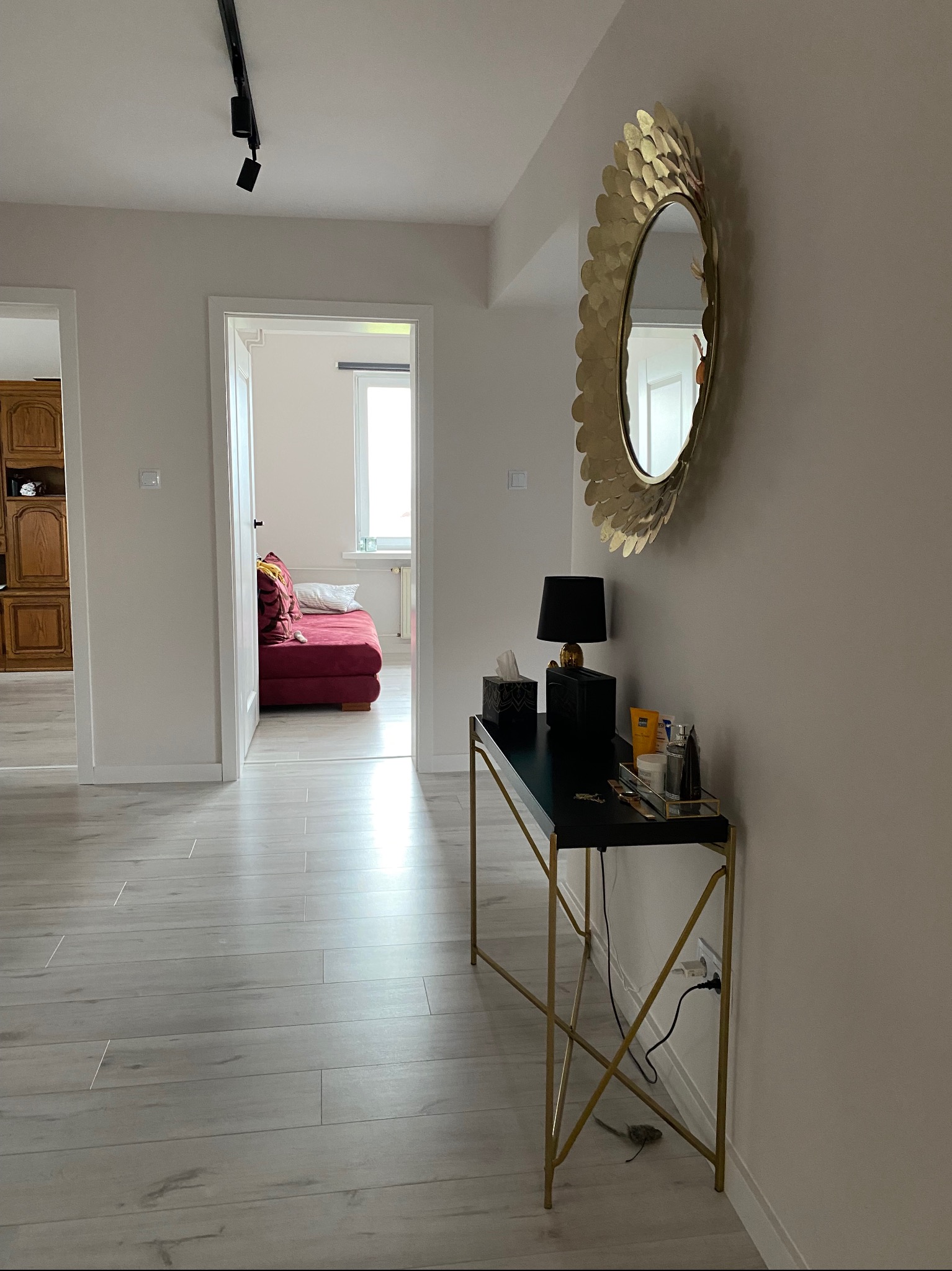 Jasne wnętrze mieszkania z drewnianą podłogą, w tle widoczny czerwony narożnik. Na ścianie dekoracyjne, złote lustro w kształcie słońca, a pod nim czarna konsola ze złotymi nogami.