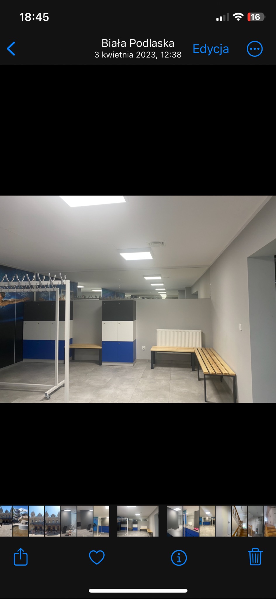 Szara szatnia z niebiesko-biało-czarnymi szafkami, drewnianymi ławkami na czarnych metalowych nogach oraz wieszakami na ubrania przy ścianie z motywem pływackim.