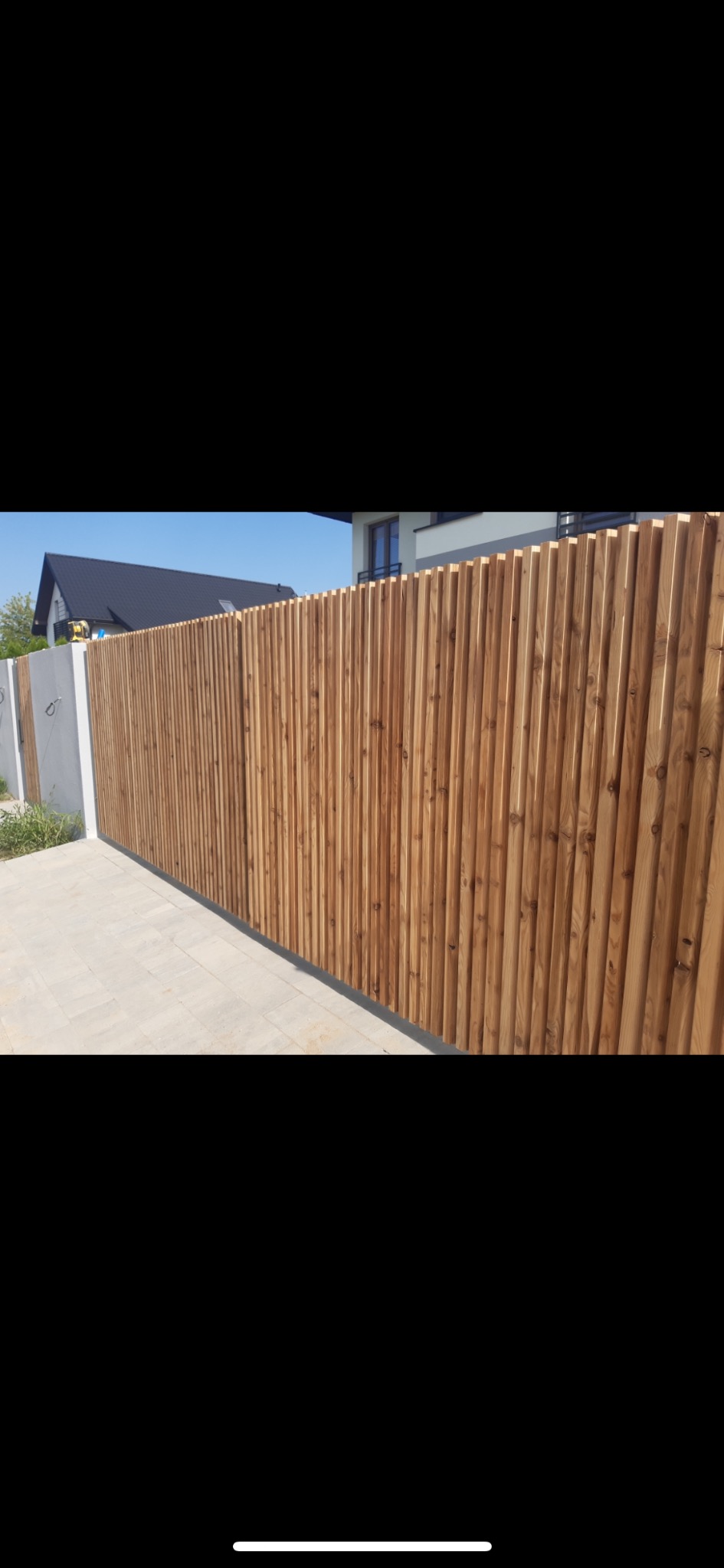 Nowoczesne ogrodzenie z pionowych desek z jasnego drewna, osadzone na betonowych słupkach, na tle domu z ciemnym dachem i błękitnego nieba.