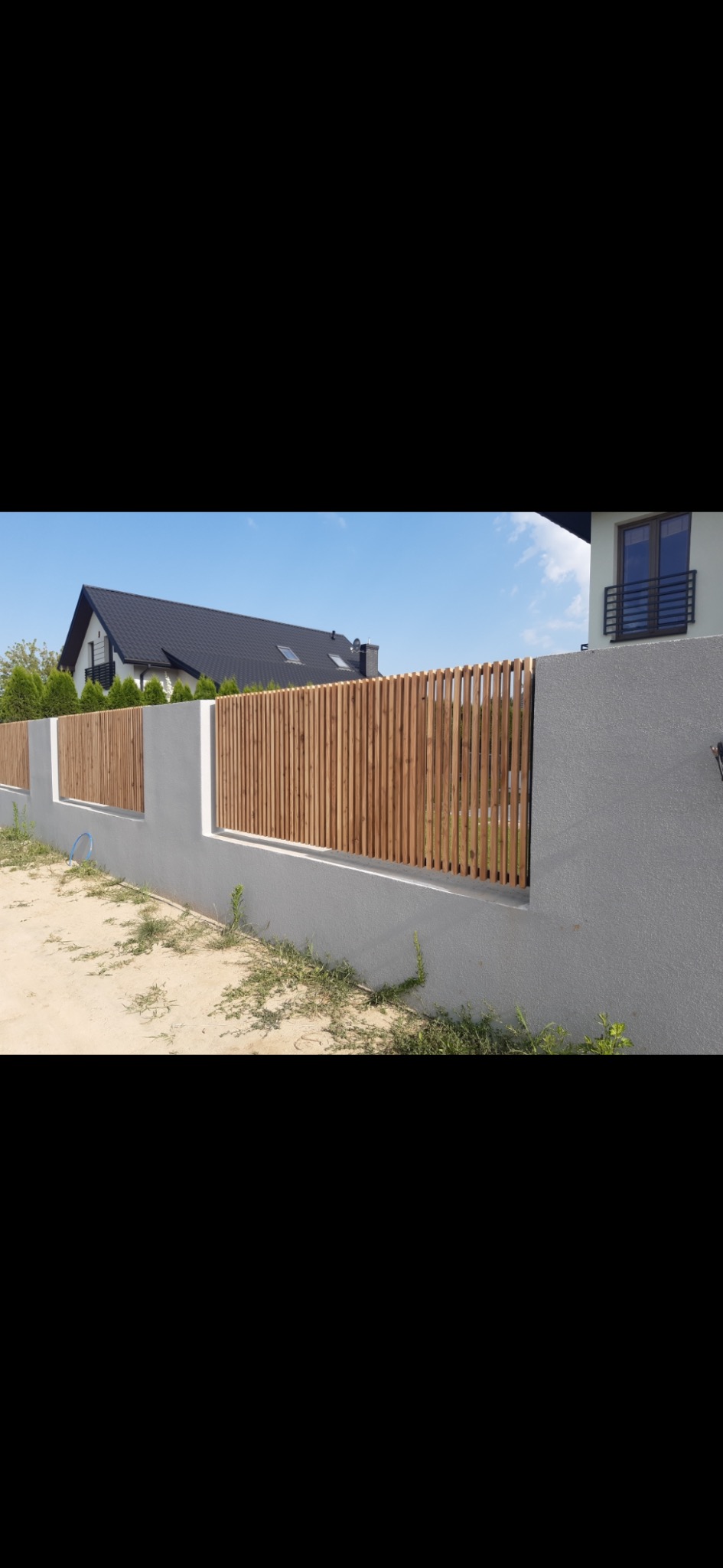 Nowoczesne ogrodzenie z drewnianych listew i szarego betonu, widok na dom z czarnym dachem w tle.
