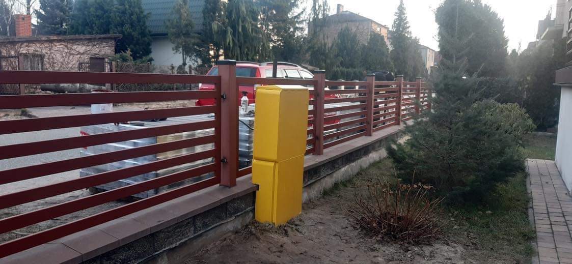 Nowoczesne, poziome ogrodzenie z brązowych listew i słupków, z żółtą skrzynką gazową przy jednym ze słupków. W tle widoczny budynek mieszkalny i zieleń.