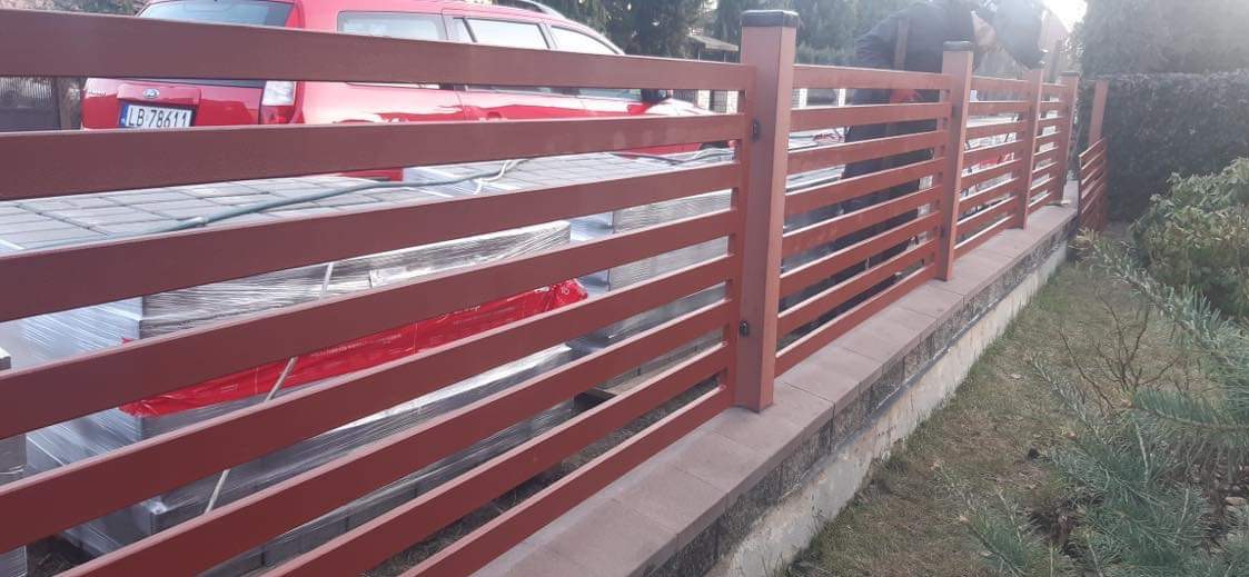 Nowoczesne ogrodzenie posesji z poziomych, brązowych elementów, widoczne zapakowane płyty chodnikowe i czerwony samochód w tle.