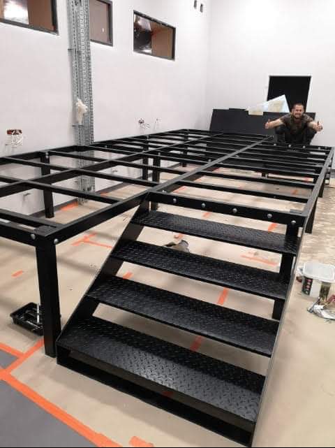 Metalowa konstrukcja pod podłogę z schodami w surowym wnętrzu biura, pomarańczowa taśma na podłodze, mężczyzna siedzący na konstrukcji.