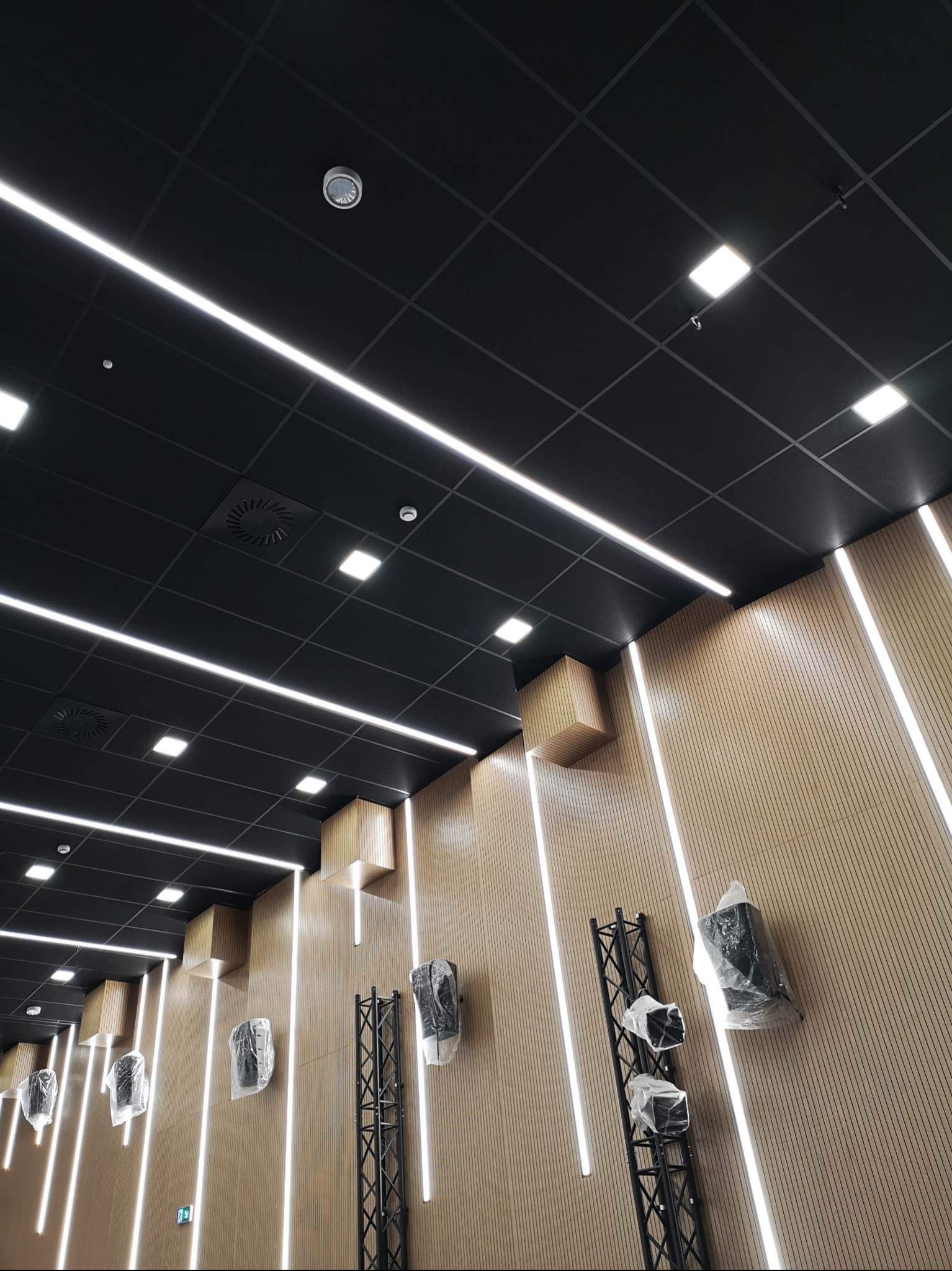 Nowoczesne oświetlenie biurowe: czarny sufit kasetonowy z liniowymi lampami LED, drewniana ściana z pionowymi panelami i elementami dekoracyjnymi, sprzęt nagłośnieniowy zabezpieczony folią.