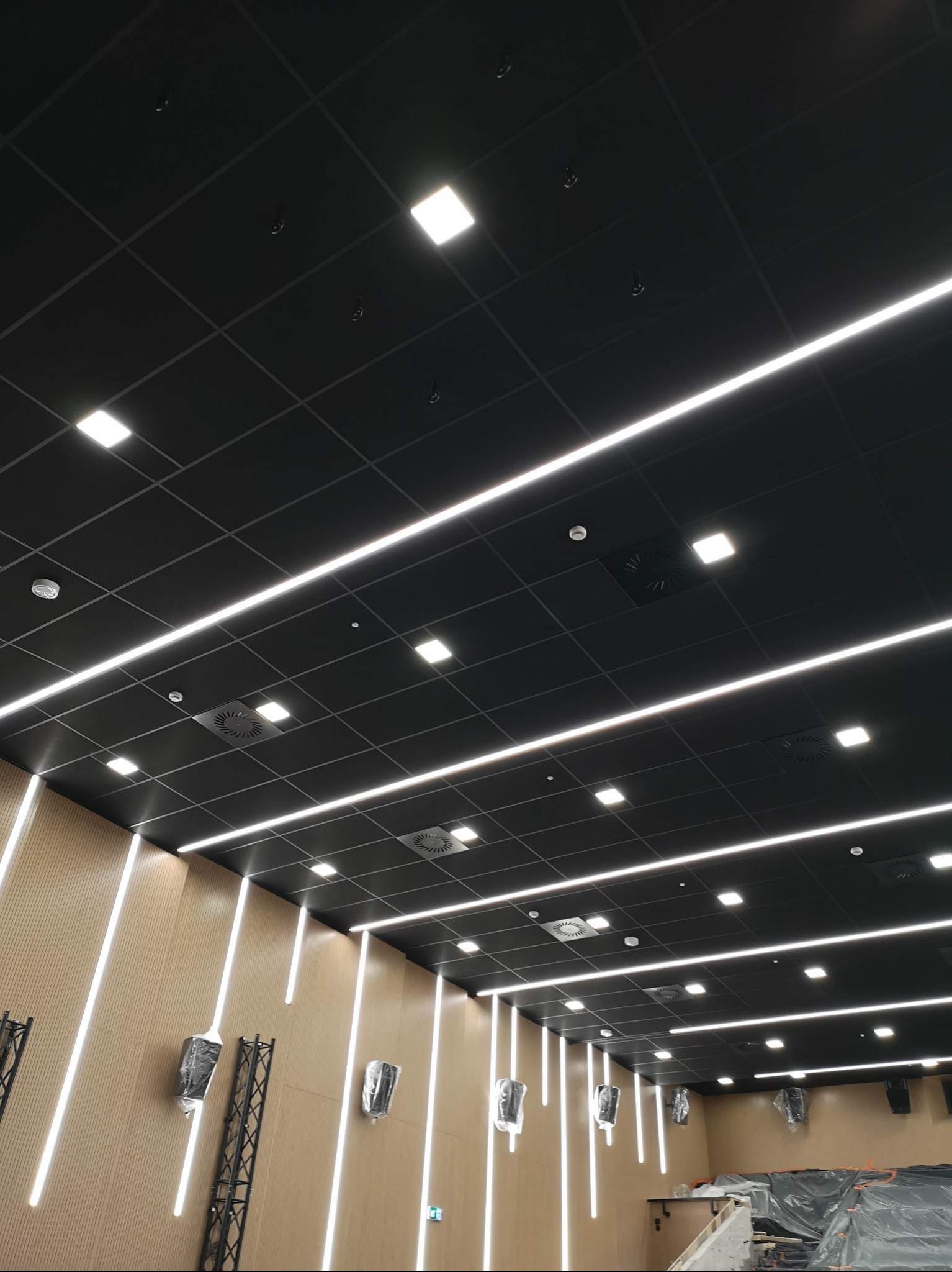 Czarny sufit podwieszany z kwadratowymi panelami, białymi liniowymi lampami LED i kratkami wentylacyjnymi, widok na ścianę z drewnopodobnych paneli z pionowymi paskami LED, elementy konstrukcyjne...