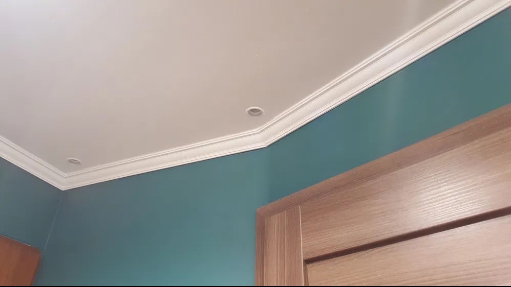 Turkusowa ściana z białym sufitem z listwą dekoracyjną i wpuszczonymi punktami świetlnymi, fragment drewnianej ościeżnicy drzwiowej w prawym dolnym rogu.