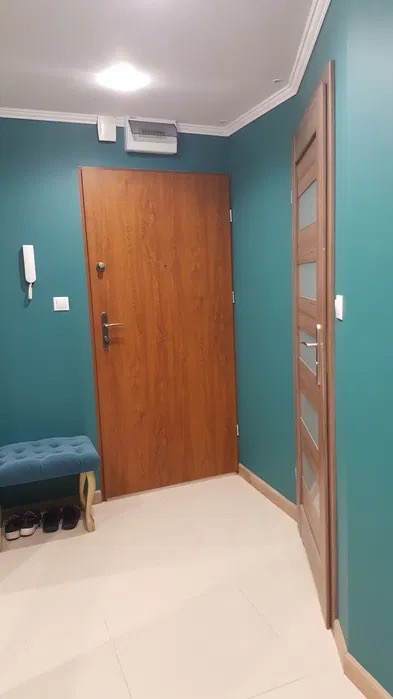 Korytarz w odcieniach turkusu z dwiema drewnianymi drzwiami: wejściowymi i wewnętrznymi z przeszkleniami, na podłodze jasne płytki, mała pufa z butami pod ścianą.