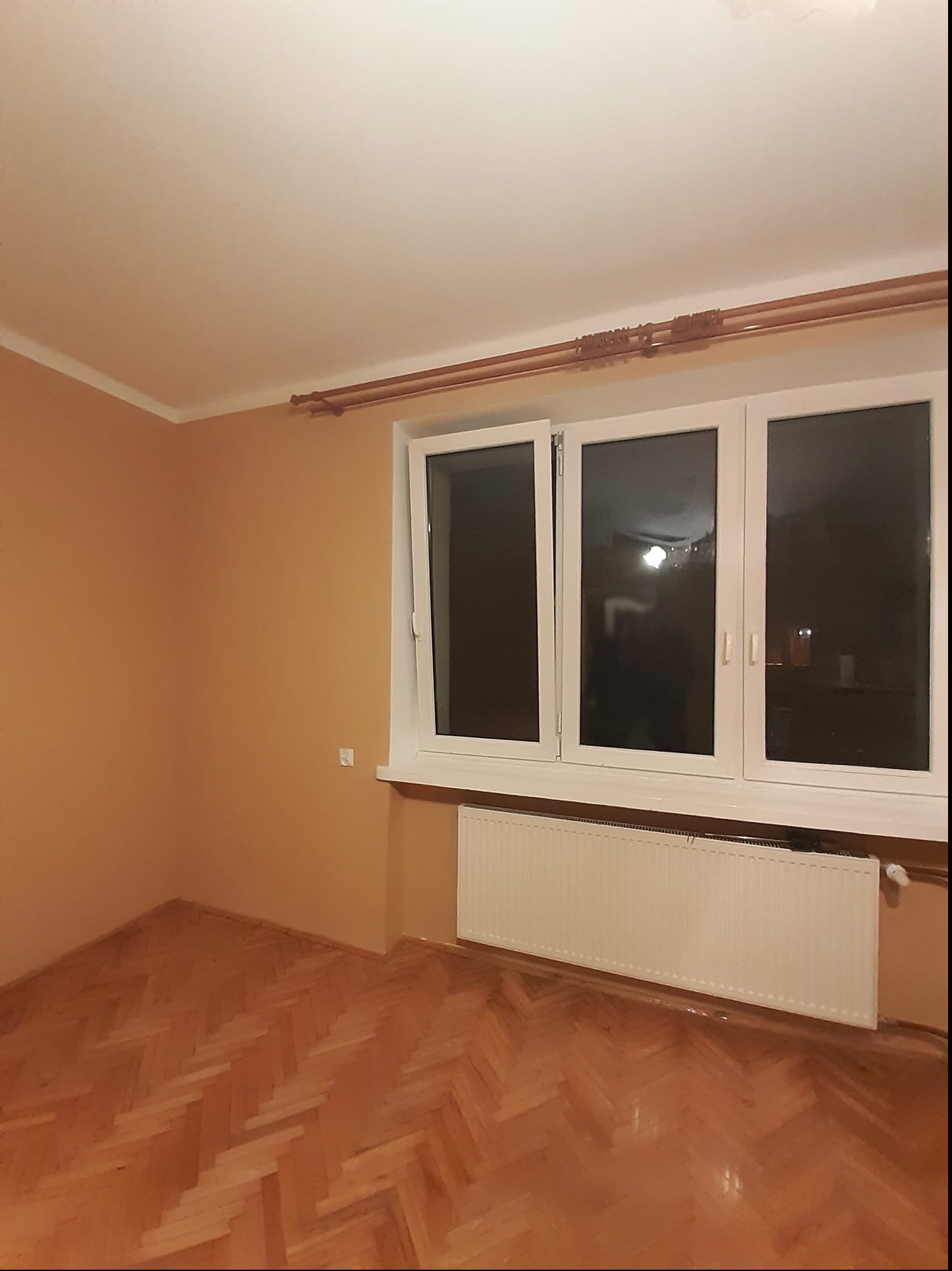 Pomalowane na ciepły odcień wnętrze pokoju z parkietem ułożonym w jodełkę, białym grzejnikiem pod trzyskrzydłowym oknem z otwartym jednym skrzydłem.