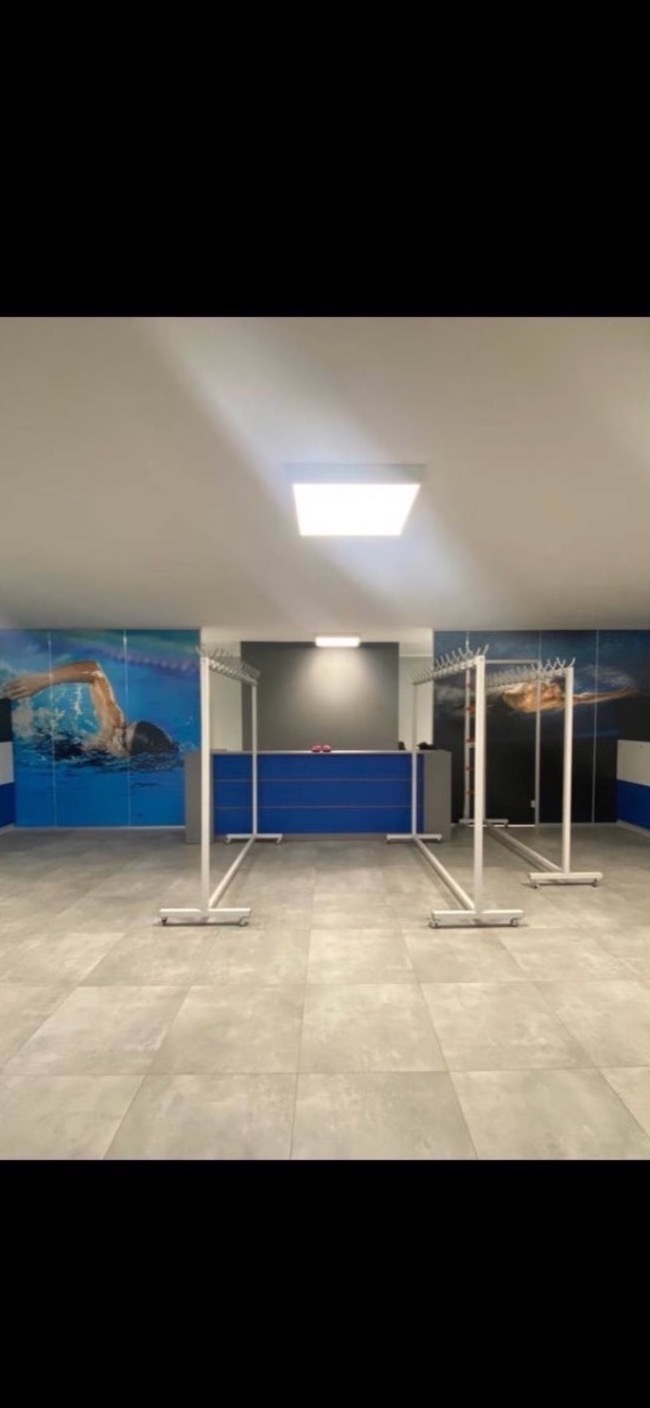 Wnętrze szatni z szaro-beżową podłogą z płytek, białe wieszaki na kółkach, niebieska lada recepcyjna i mural z pływakiem na ścianie.