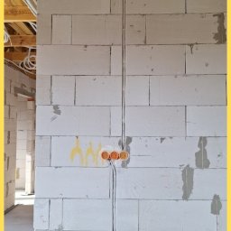 Srokowski Instalacje elektryczne - Ściana z betonu komórkowego z poprowadzonymi przewodami elektrycznymi w peszlach i zamontowanymi puszkami instalacyjnymi. Widoczne drewniane elementy konstrukcji dachu.