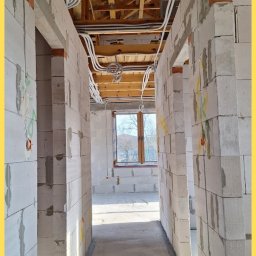 Srokowski Instalacje elektryczne - Wnętrze w trakcie budowy z widoczną instalacją elektryczną pod sufitem, kable w peszlach. Ściany z białych bloczków, widoczne otwory na gniazdka. Surowy stan.