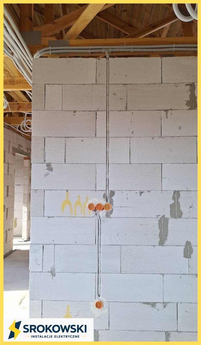 Ściana z betonu komórkowego z poprowadzonymi przewodami elektrycznymi w peszlach i zamontowanymi puszkami instalacyjnymi. Widoczne drewniane elementy konstrukcji dachu.