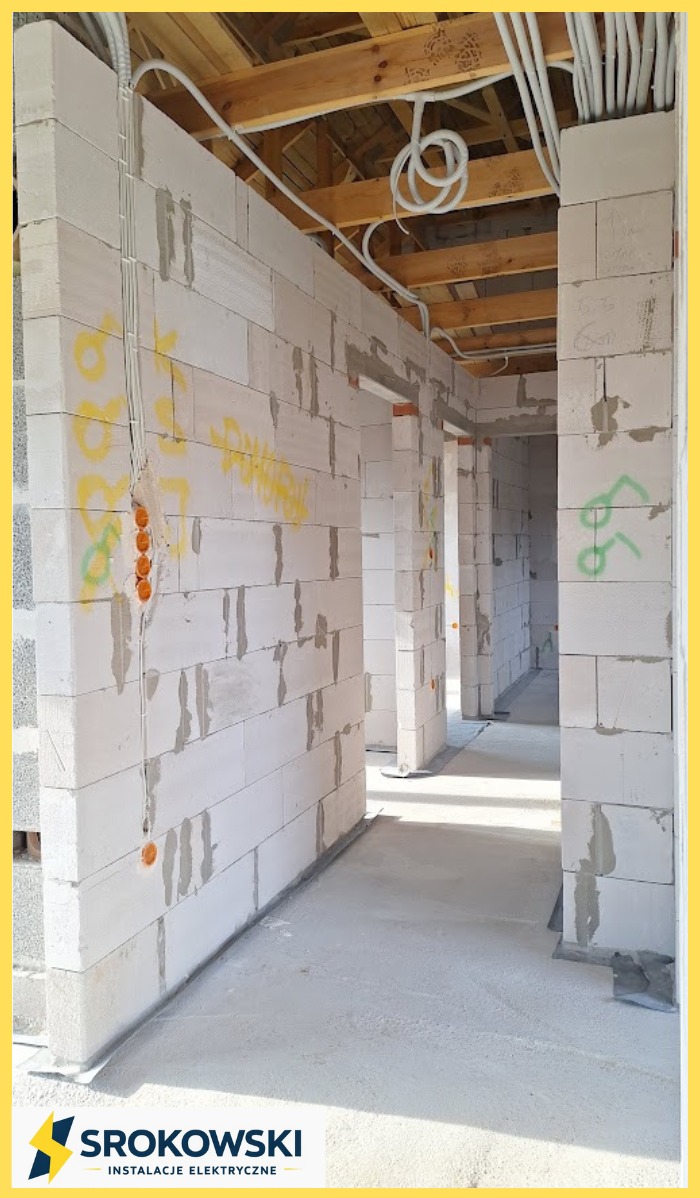 Instalacja elektryczna w surowym budynku z białego bloczka: kable prowadzone po ścianach i pod sufitem, widoczne puszki instalacyjne. Etap prac przed tynkowaniem.