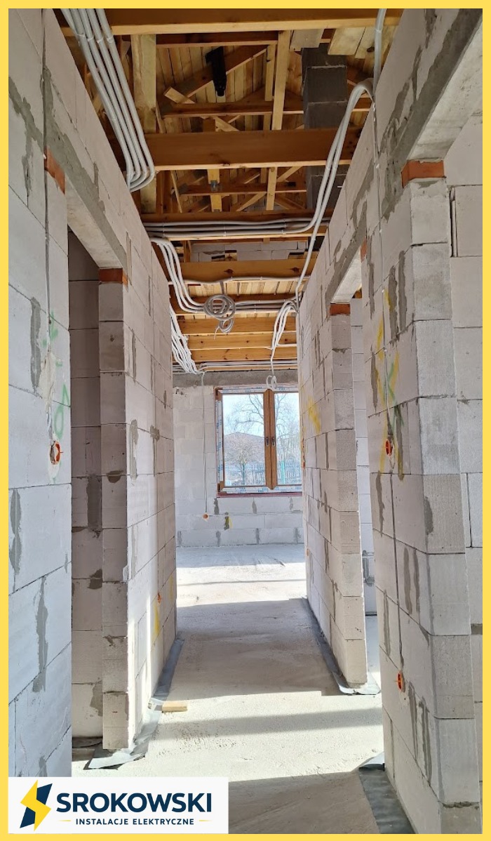 Wnętrze w trakcie budowy z widoczną instalacją elektryczną pod sufitem, kable w peszlach. Ściany z białych bloczków, widoczne otwory na gniazdka. Surowy stan.