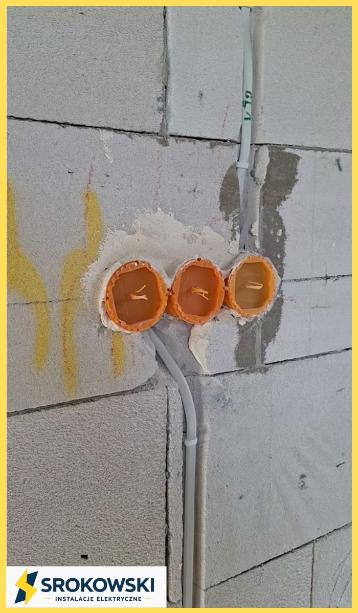 Instalacja elektryczna w trakcie montażu: trzy pomarańczowe puszki podtynkowe osadzone w ścianie z betonu komórkowego, z widocznymi przewodami i peszlami.