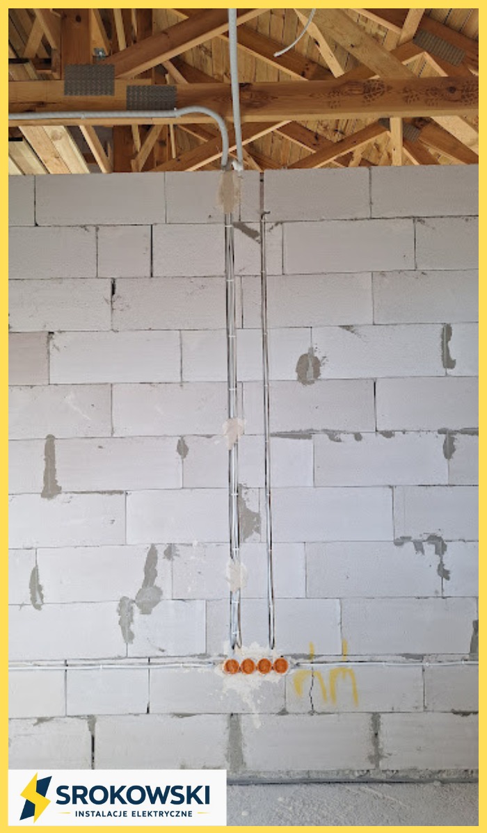 Ściana z betonu komórkowego z zamontowanymi rurami instalacji elektrycznej i puszkami pod gniazdka. Widoczne drewniane elementy konstrukcji dachu.