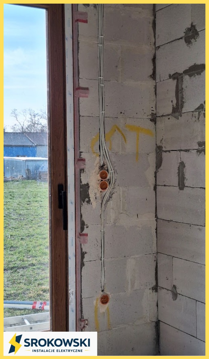 Ściana z instalacją elektryczną w trakcie montażu, widoczne kable w peszlach i puszki pod gniazdka, surowy mur z betonu komórkowego, okno z widokiem na zewnątrz.