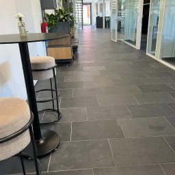 MZ - SYSTEM - MARIUSZ ZALEWSKI - Wnętrze biura z czarną podłogą z płytek, stolikiem barowym z kwiatem i krzesłami. Widok na korytarz z przeszklonymi ścianami i roślinami w tle.