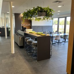 MZ - SYSTEM - MARIUSZ ZALEWSKI - Nowoczesna przestrzeń biurowa z wyspą kuchenną, barowymi stołkami i roślinami zwisającymi z sufitu. Widoczne szklanki i przekąski na blacie.