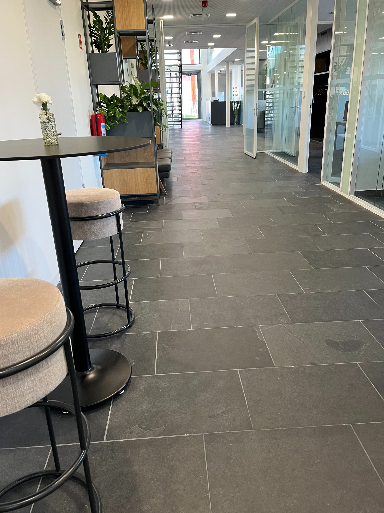 Wnętrze biura z czarną podłogą z płytek, stolikiem barowym z kwiatem i krzesłami. Widok na korytarz z przeszklonymi ścianami i roślinami w tle.