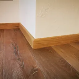 Fragment podłogi z ciemnego drewna z widocznymi słojami, wykończonej jasnymi listwami przypodłogowymi przy białej ścianie z fakturą.